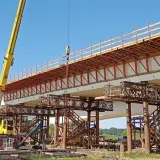 Budowa mostu metalowego w toku. Żółty dźwig podnosi elementy konstrukcji nad betonowymi podporami.