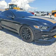 Czarny Mustang GT 2015 z żółtymi pasami na masce i zderzaku, zaparkowany na żwirowisku.