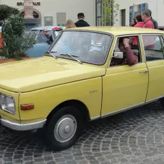 Żółty samochód Wartburg z lat 80. stoi na brukowanej ulicy. Kierowca opiera rękę na drzwiach.