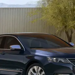 Ciemnoniebieski Chevrolet Impala z chromowanymi detalami stoi przed nowoczesnym ogrodzeniem i drzewem.
