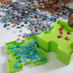Dwie zielone tacki pomagają w układaniu puzzli. Jedna z nich zawiera niebieskie elementy, druga czerwone. Na stole leżą setki rozsypanych kawałków.