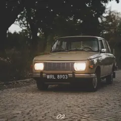 Beżowy Wartburg 353 z włączonymi światłami na brukowanej drodze, otoczony jesienną zielenią.