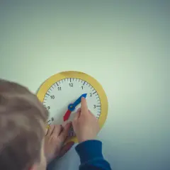Dziecko bawi się zegarem, który wygląda jak zegar chodzący do tyłu. Wskazówka godzinowa jest na 2, a minutowa na 10.