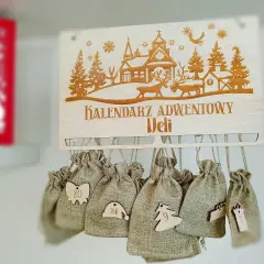 Drewniany kalendarz adwentowy z wygrawerowaną zimową scenką i zawieszonymi woreczkami z numerami.