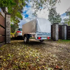 Przyczepka drewniana z plandeką, gotowa do drogi, zaparkowana przy bramie.