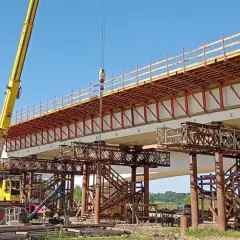Budowa mostu metalowego w toku. Żółty dźwig podnosi elementy konstrukcji nad betonowymi podporami.