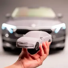 Dłoń trzyma szary model samochodu 3d na tle prawdziwego samochodu z zapalonymi światłami.