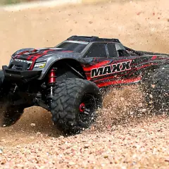 Czarny i czerwony samochód monster truck z napisem "MAXX" pędzi po piaszczystym terenie, wzbijając tumany kurzu.