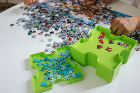 Dwie zielone tacki pomagają w układaniu puzzli. Jedna z nich zawiera niebieskie elementy, druga czerwone. Na stole leżą setki rozsypanych kawałków.
