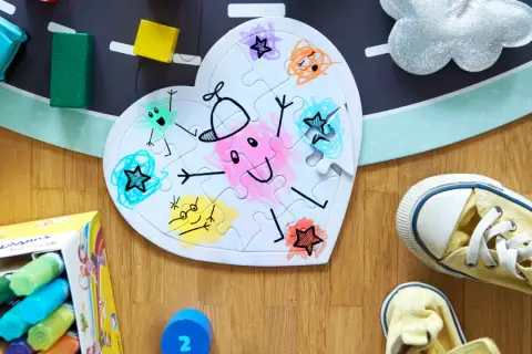 Serce z puzzli do malowania, ozdobione kolorowymi rysunkami postaci i gwiazdek. Obok leżą kredki i zabawki.