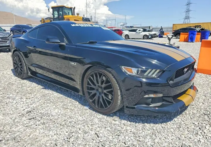 Czarny Mustang GT 2015 z żółtymi pasami na masce i zderzaku, zaparkowany na żwirowisku.