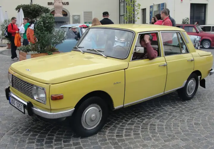 Żółty samochód Wartburg z lat 80. stoi na brukowanej ulicy. Kierowca opiera rękę na drzwiach.