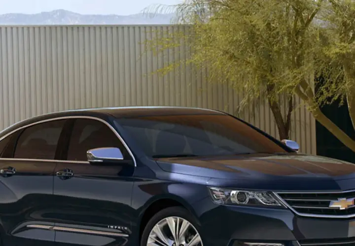Ciemnoniebieski Chevrolet Impala z chromowanymi detalami stoi przed nowoczesnym ogrodzeniem i drzewem.