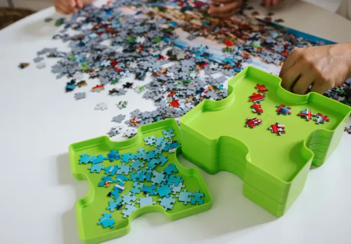 Dwie zielone tacki pomagają w układaniu puzzli. Jedna z nich zawiera niebieskie elementy, druga czerwone. Na stole leżą setki rozsypanych kawałków.