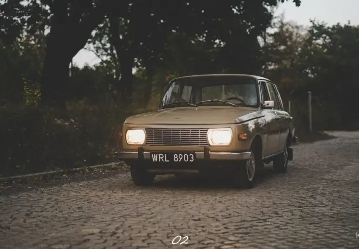 Beżowy Wartburg 353 z włączonymi światłami na brukowanej drodze, otoczony jesienną zielenią.