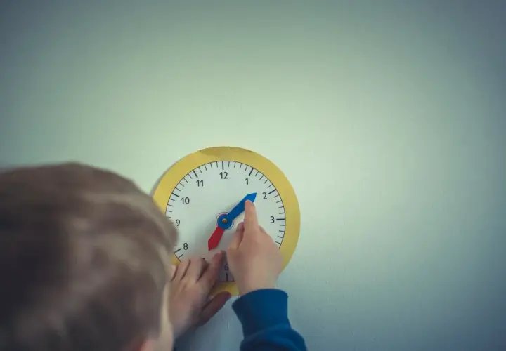 Dziecko bawi się zegarem, który wygląda jak zegar chodzący do tyłu. Wskazówka godzinowa jest na 2, a minutowa na 10.