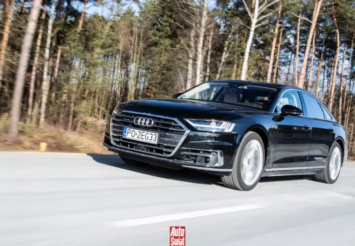 Czarne Audi A8 sunie po drodze wśród drzew. To jak układanie puzzli z luksusu i dynamiki.