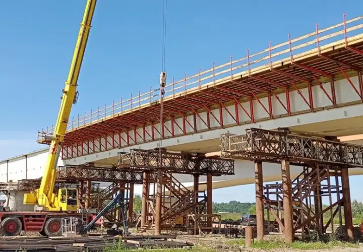 Budowa mostu metalowego w toku. Żółty dźwig podnosi elementy konstrukcji nad betonowymi podporami.