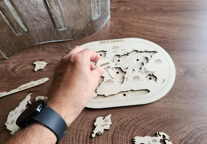 Dłoń układa drewniane puzzle kontynenty na mapie świata. Antarktyda i Europa czekają na swoje miejsce.