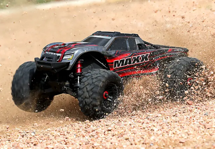 Czarny i czerwony samochód monster truck z napisem "MAXX" pędzi po piaszczystym terenie, wzbijając tumany kurzu.