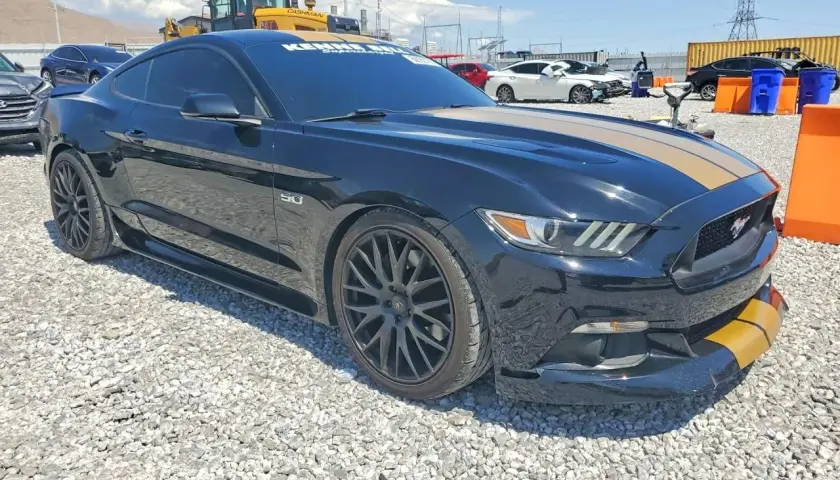 Czarny Mustang GT 2015 z żółtymi pasami na masce i zderzaku, zaparkowany na żwirowisku.