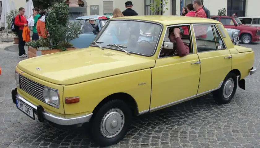 Żółty samochód Wartburg z lat 80. stoi na brukowanej ulicy. Kierowca opiera rękę na drzwiach.
