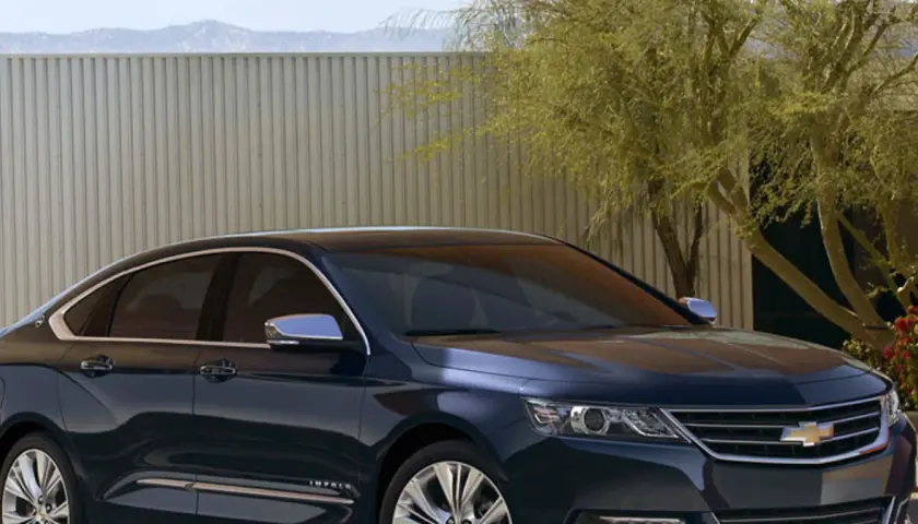 Ciemnoniebieski Chevrolet Impala z chromowanymi detalami stoi przed nowoczesnym ogrodzeniem i drzewem.