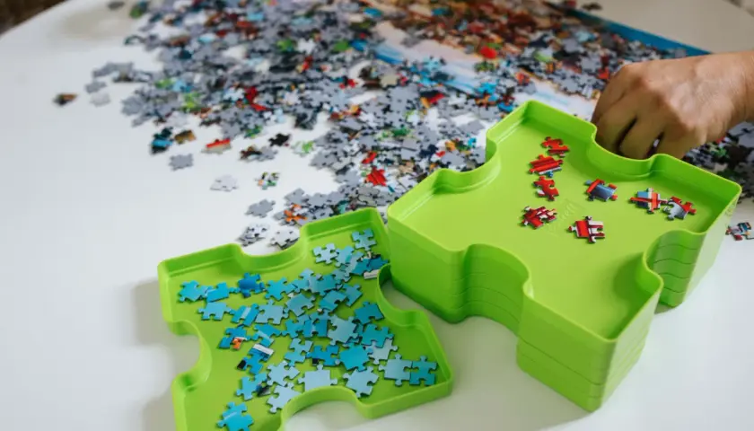 Dwie zielone tacki pomagają w układaniu puzzli. Jedna z nich zawiera niebieskie elementy, druga czerwone. Na stole leżą setki rozsypanych kawałków.