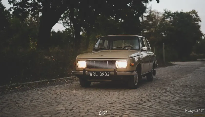 Beżowy Wartburg 353 z włączonymi światłami na brukowanej drodze, otoczony jesienną zielenią.