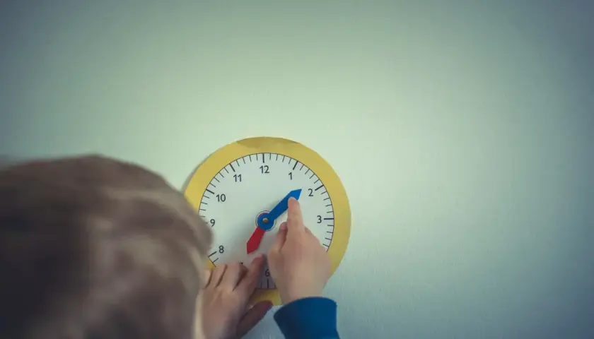 Dziecko bawi się zegarem, który wygląda jak zegar chodzący do tyłu. Wskazówka godzinowa jest na 2, a minutowa na 10.
