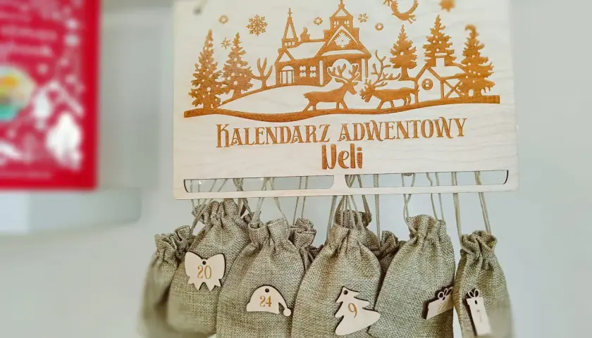 Drewniany kalendarz adwentowy z wygrawerowaną zimową scenką i zawieszonymi woreczkami z numerami.