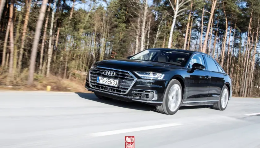 Czarne Audi A8 sunie po drodze wśród drzew. To jak układanie puzzli z luksusu i dynamiki.