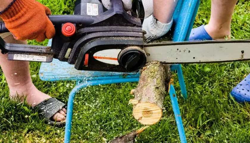 Ręce w rękawiczkach trzymają piłę łańcuchową, która tnie drewno. W tle widać nogi i trawę. Bączek do drewna jest w użyciu.