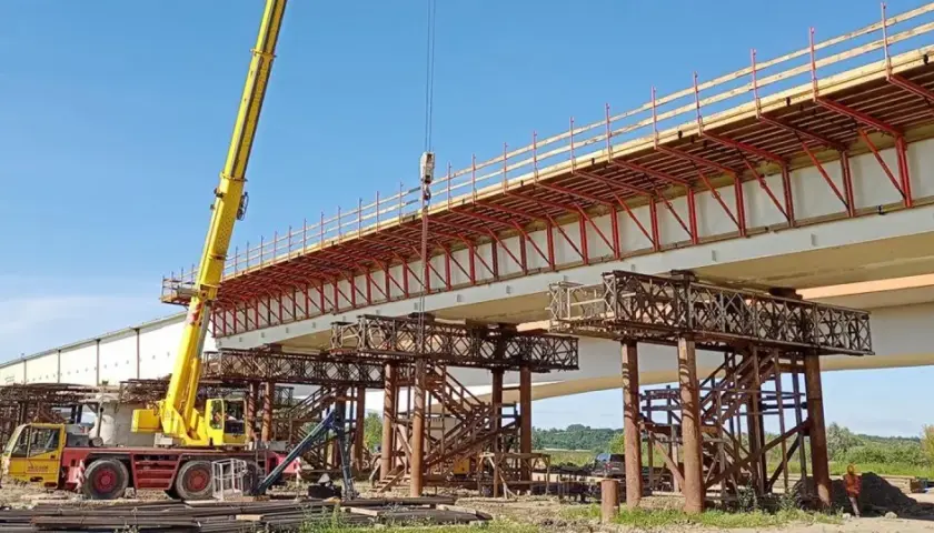 Budowa mostu metalowego w toku. Żółty dźwig podnosi elementy konstrukcji nad betonowymi podporami.