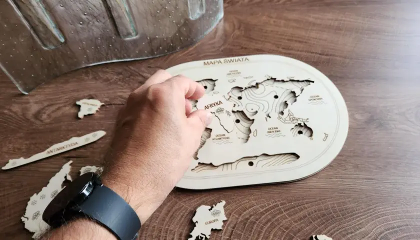 Dłoń układa drewniane puzzle kontynenty na mapie świata. Antarktyda i Europa czekają na swoje miejsce.
