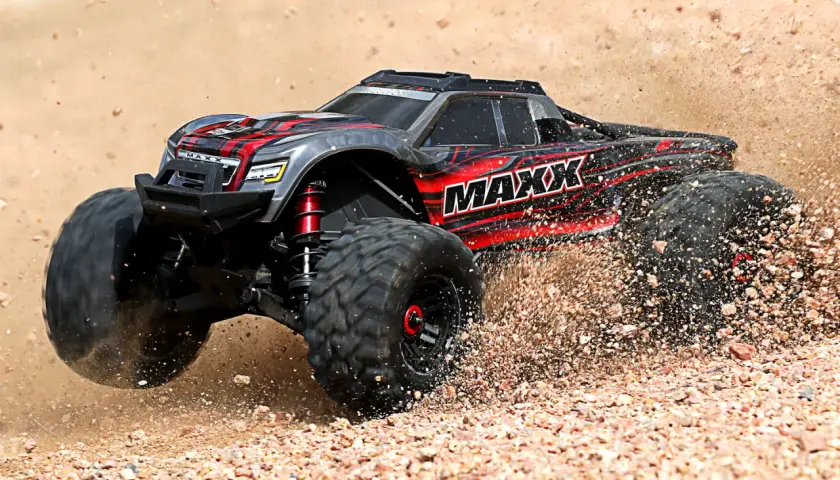 Czarny i czerwony samochód monster truck z napisem "MAXX" pędzi po piaszczystym terenie, wzbijając tumany kurzu.