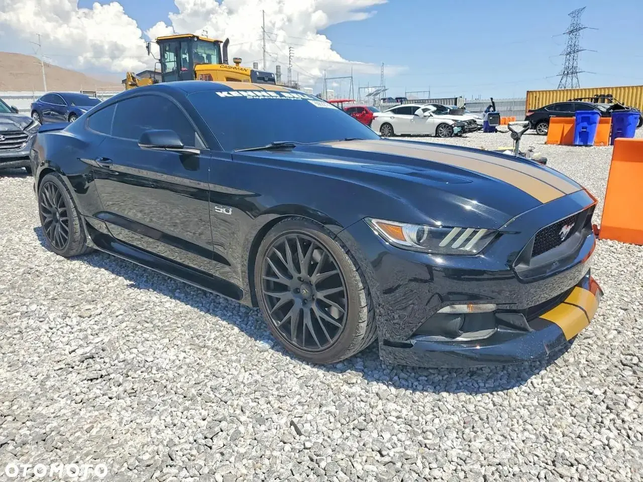 Czarny Mustang GT 2015 z żółtymi pasami na masce i zderzaku, zaparkowany na żwirowisku.