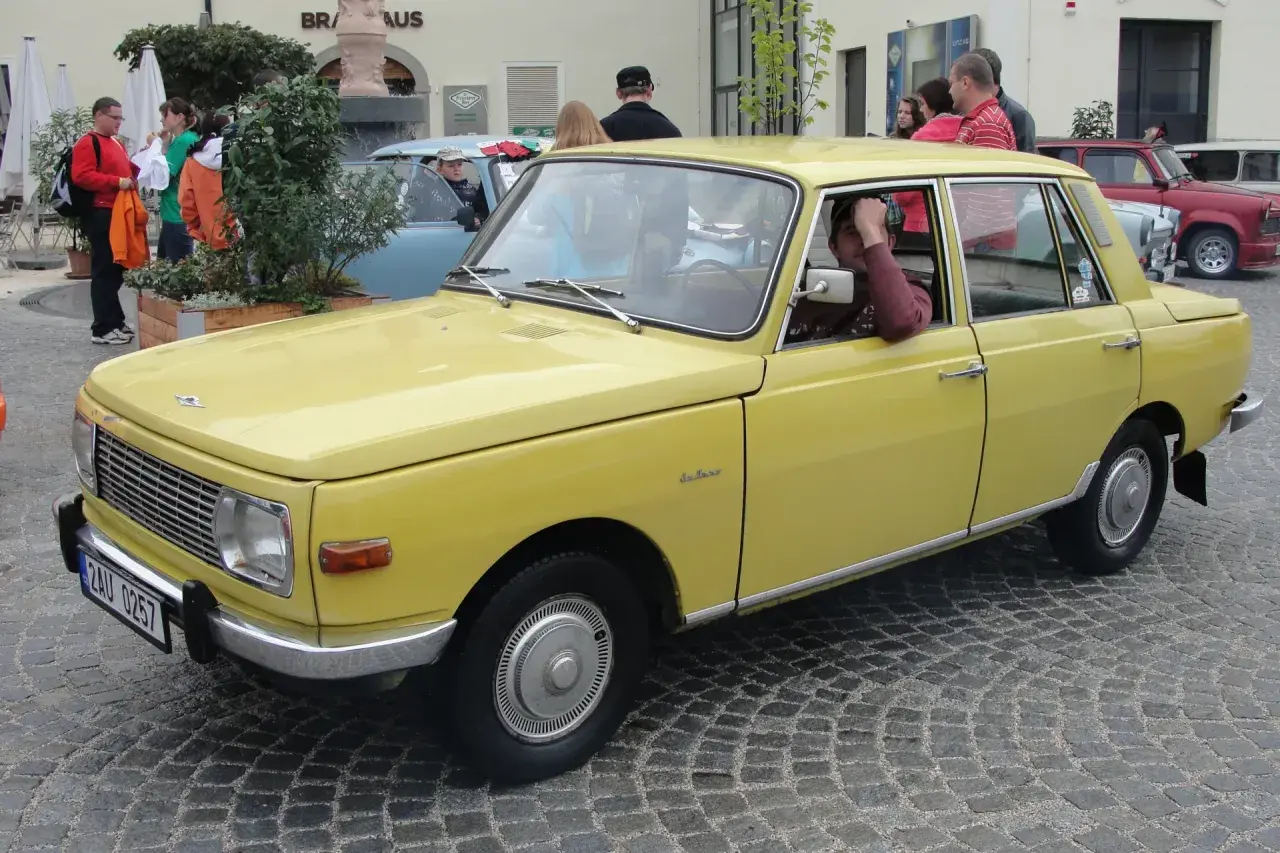 Żółty samochód Wartburg z lat 80. stoi na brukowanej ulicy. Kierowca opiera rękę na drzwiach.