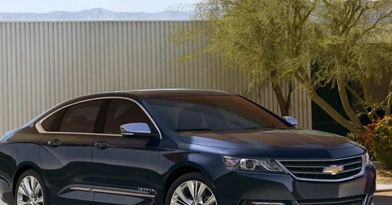 Ciemnoniebieski Chevrolet Impala z chromowanymi detalami stoi przed nowoczesnym ogrodzeniem i drzewem.