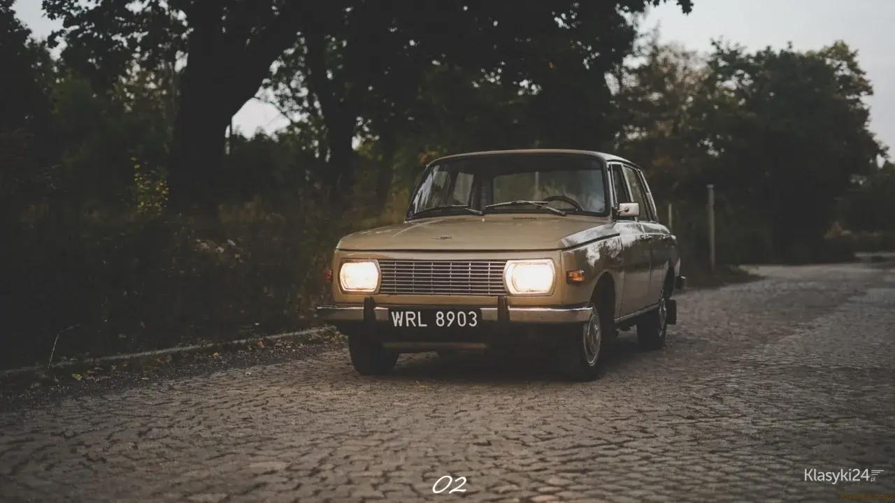 Beżowy Wartburg 353 z włączonymi światłami na brukowanej drodze, otoczony jesienną zielenią.