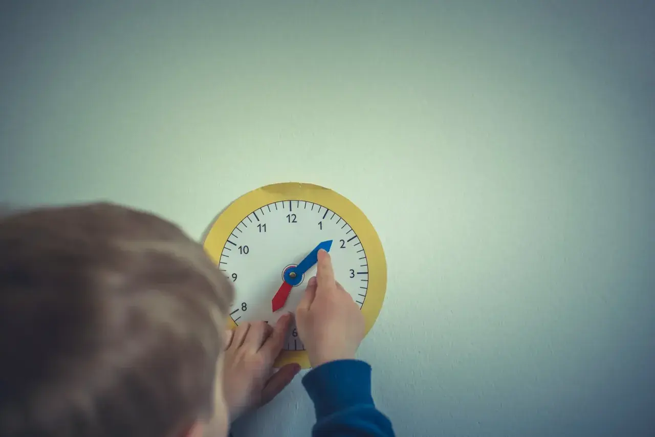 Dziecko bawi się zegarem, który wygląda jak zegar chodzący do tyłu. Wskazówka godzinowa jest na 2, a minutowa na 10.