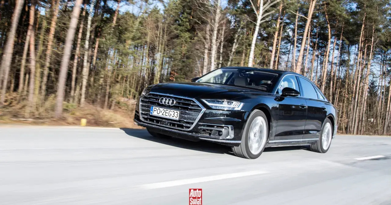 Czarne Audi A8 sunie po drodze wśród drzew. To jak układanie puzzli z luksusu i dynamiki.