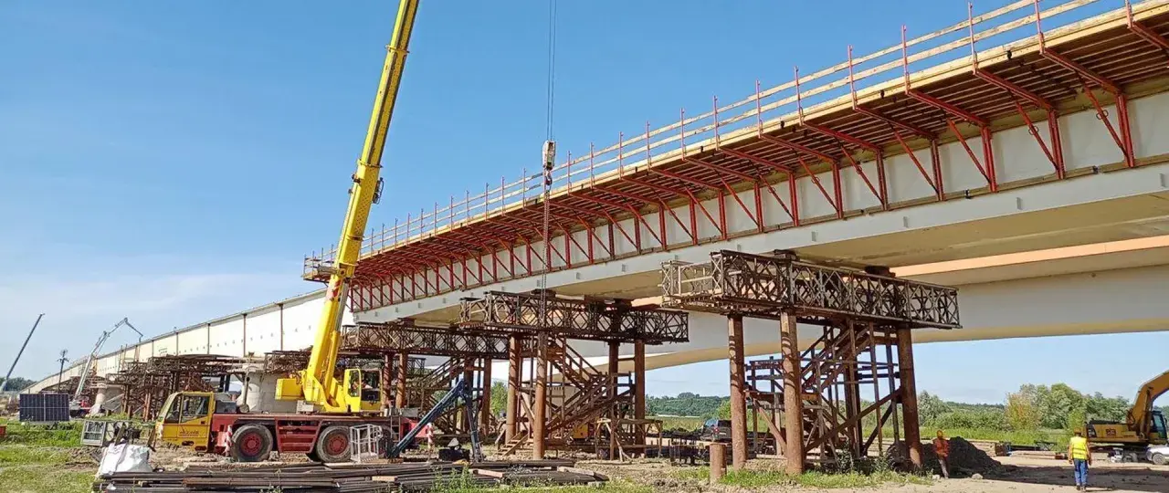 Budowa mostu metalowego w toku. Żółty dźwig podnosi elementy konstrukcji nad betonowymi podporami.