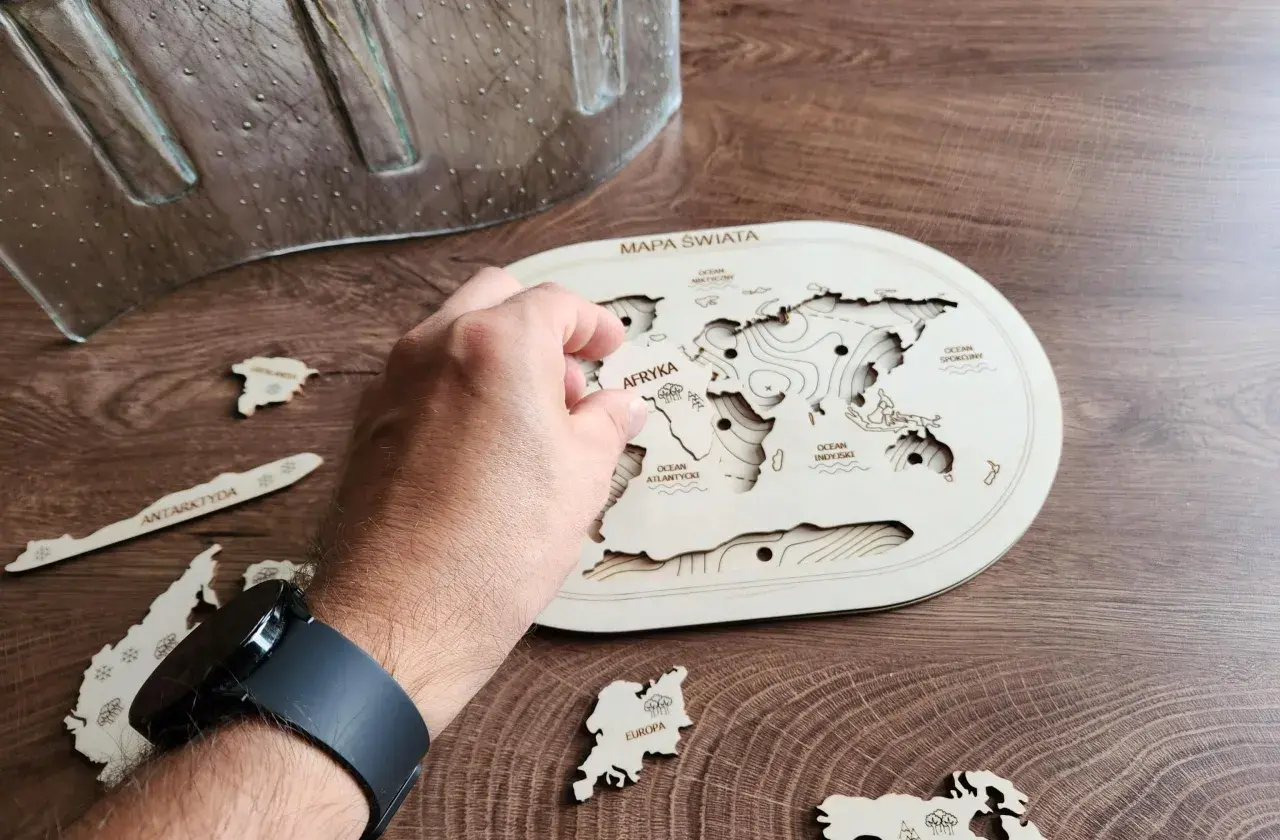 Dłoń układa drewniane puzzle kontynenty na mapie świata. Antarktyda i Europa czekają na swoje miejsce.