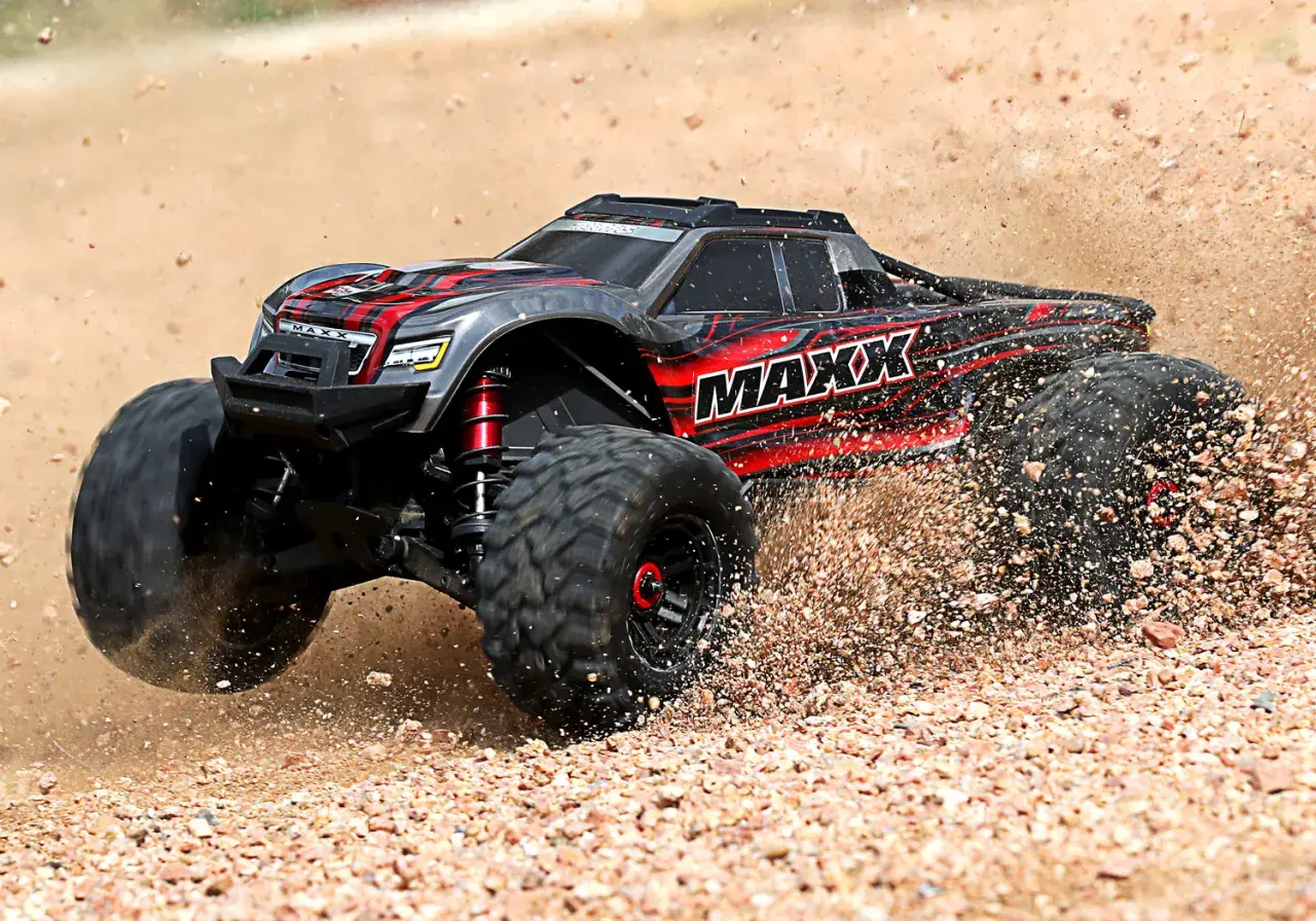 Czarny i czerwony samochód monster truck z napisem "MAXX" pędzi po piaszczystym terenie, wzbijając tumany kurzu.