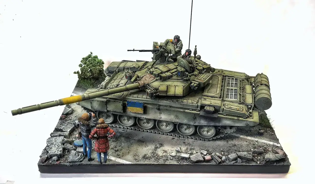 Model T-90 tank z załogą i ukraińską flagą, obok niego dwie postacie obserwują go na tle zniszczeń.