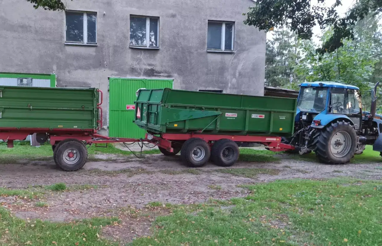 Niebieski traktor ciągnie dwa zielone, metalowe wozy do ciągnika, gotowe do pracy na gospodarstwie.