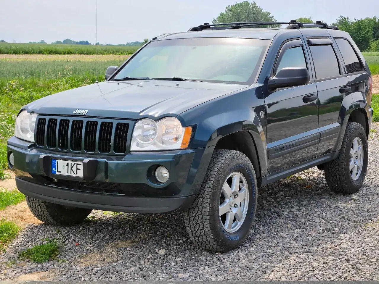Ciemnozielony Jeep Grand Cherokee, jeden z popularnych jeep modele, zaparkowany na żwirowej drodze, z polem w tle.