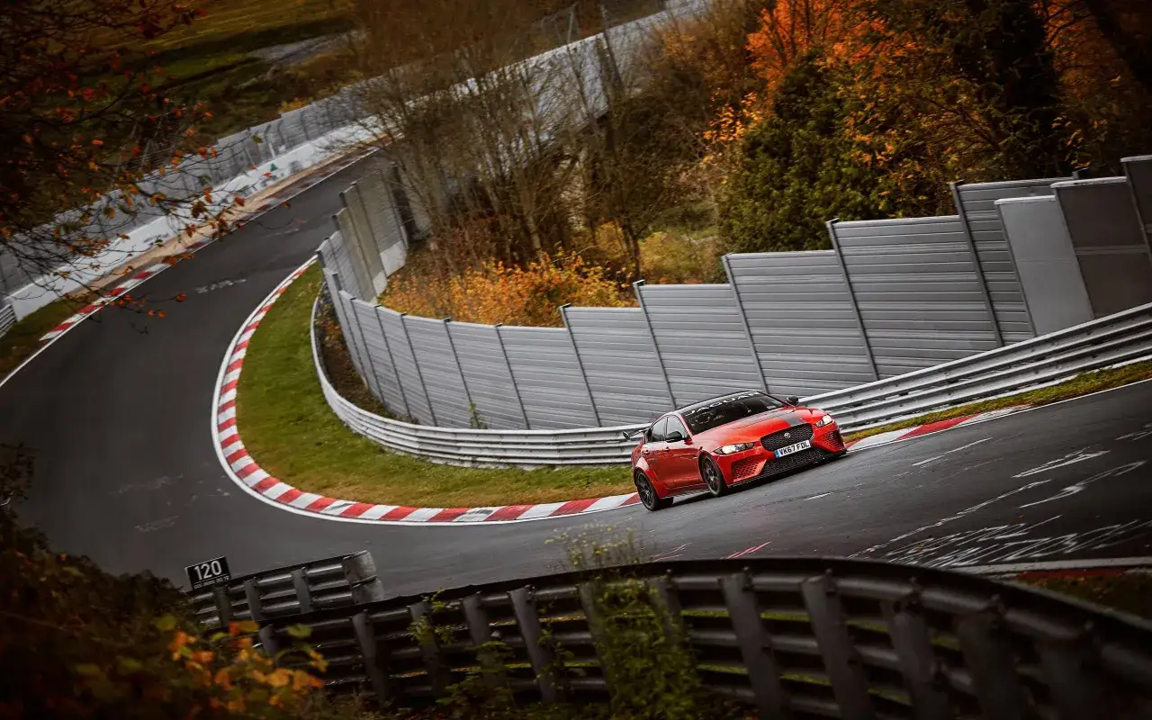 Czerwony Jaguar XE SV Project 8 pokonuje zakręt na torze wyścigowym, otoczony jesienną roślinnością i barierami ochronnymi.
