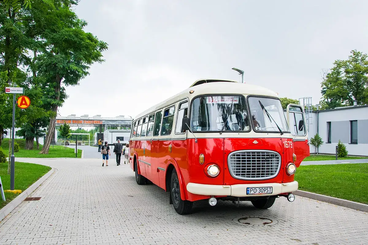Czerwony autobus og&oacute;rek z kremowym dachem, z otwartymi drzwiami, stoi na chodniku. W tle widać ludzi i budynki.